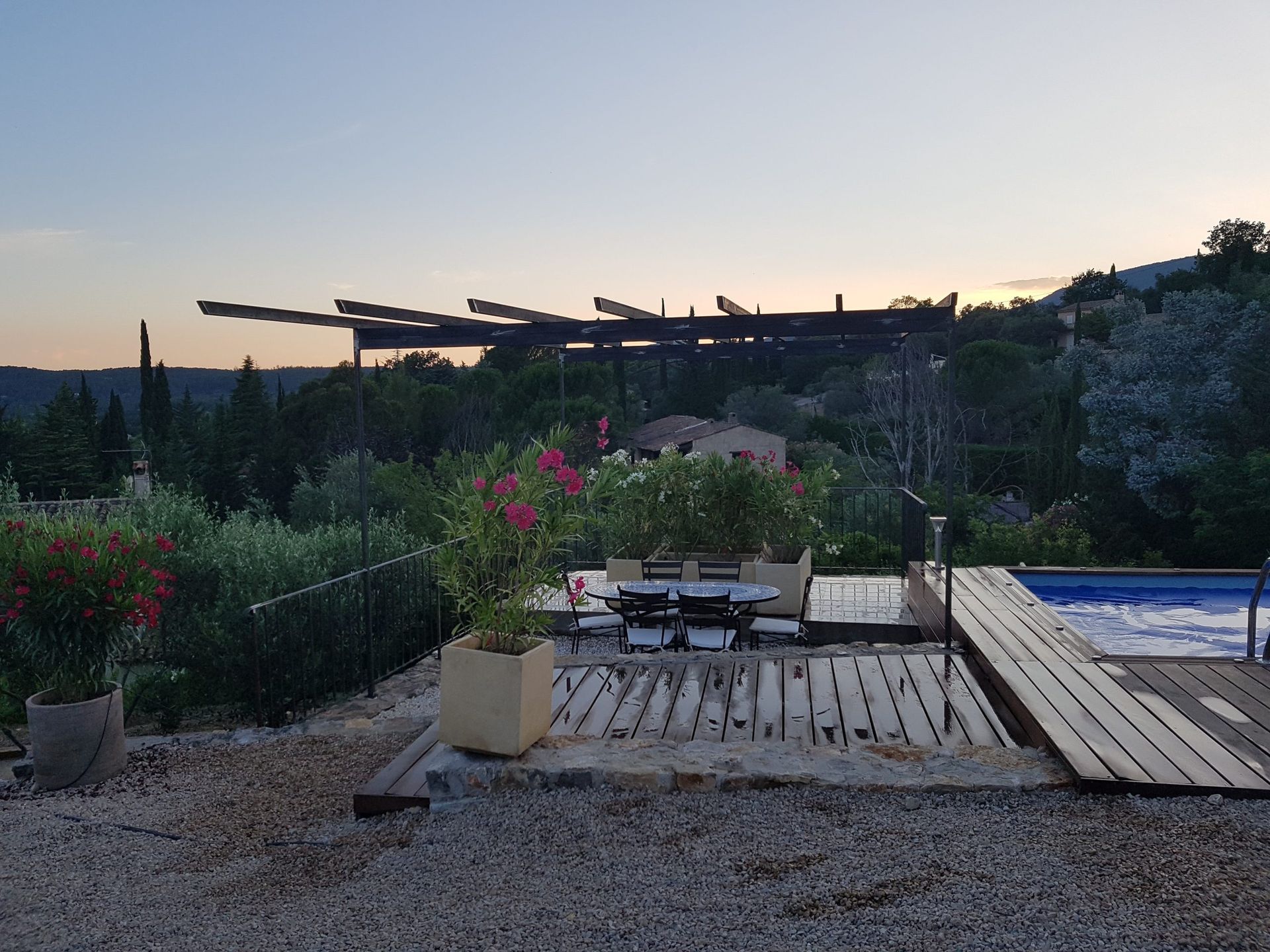 Terrasse extérieure avec plancher en bois, table et chaises, fleurs et piscine au coucher du soleil.