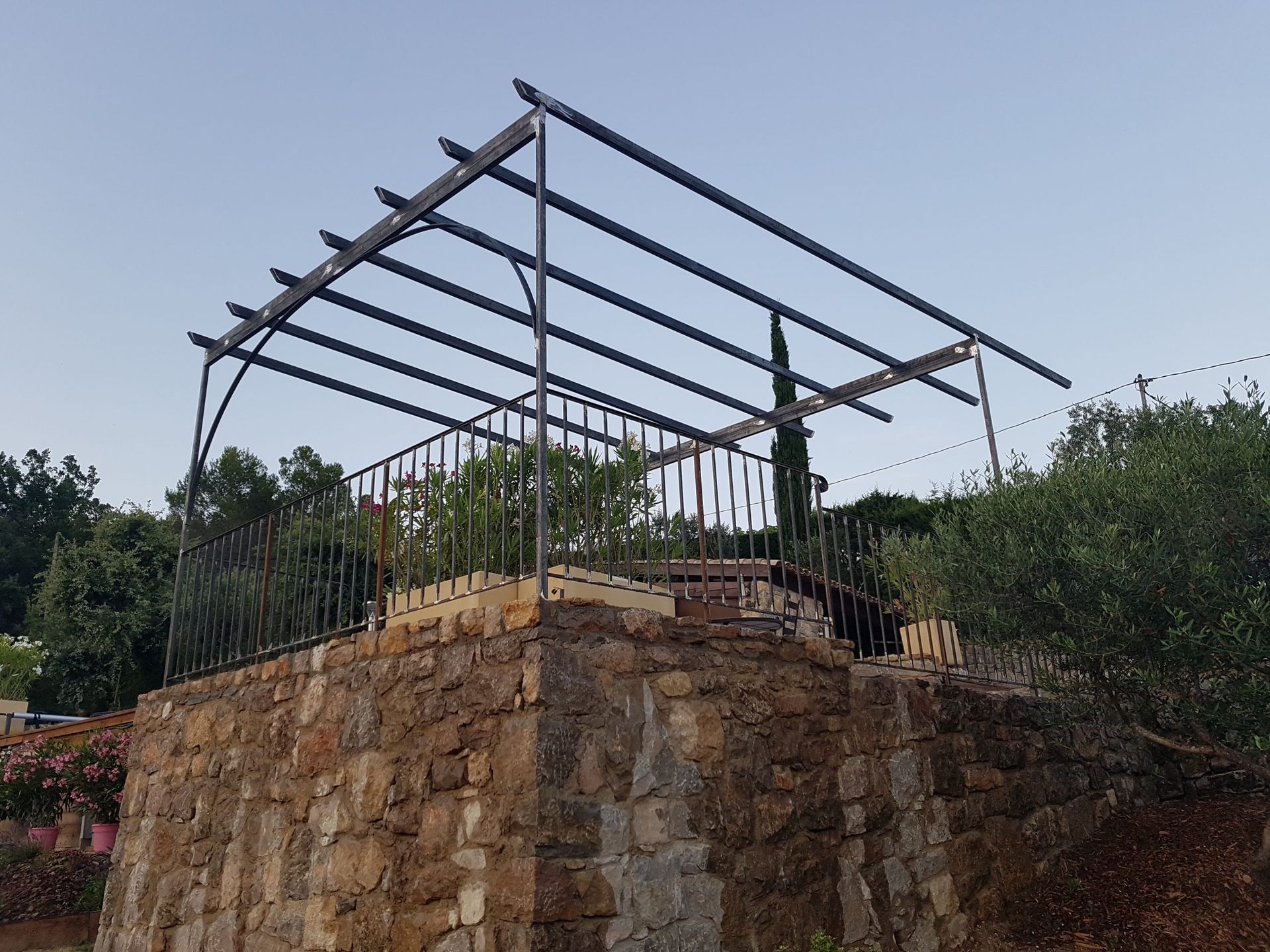 Terrasse en pierre avec une pergola en métal et verdure environnante sous un ciel dégagé