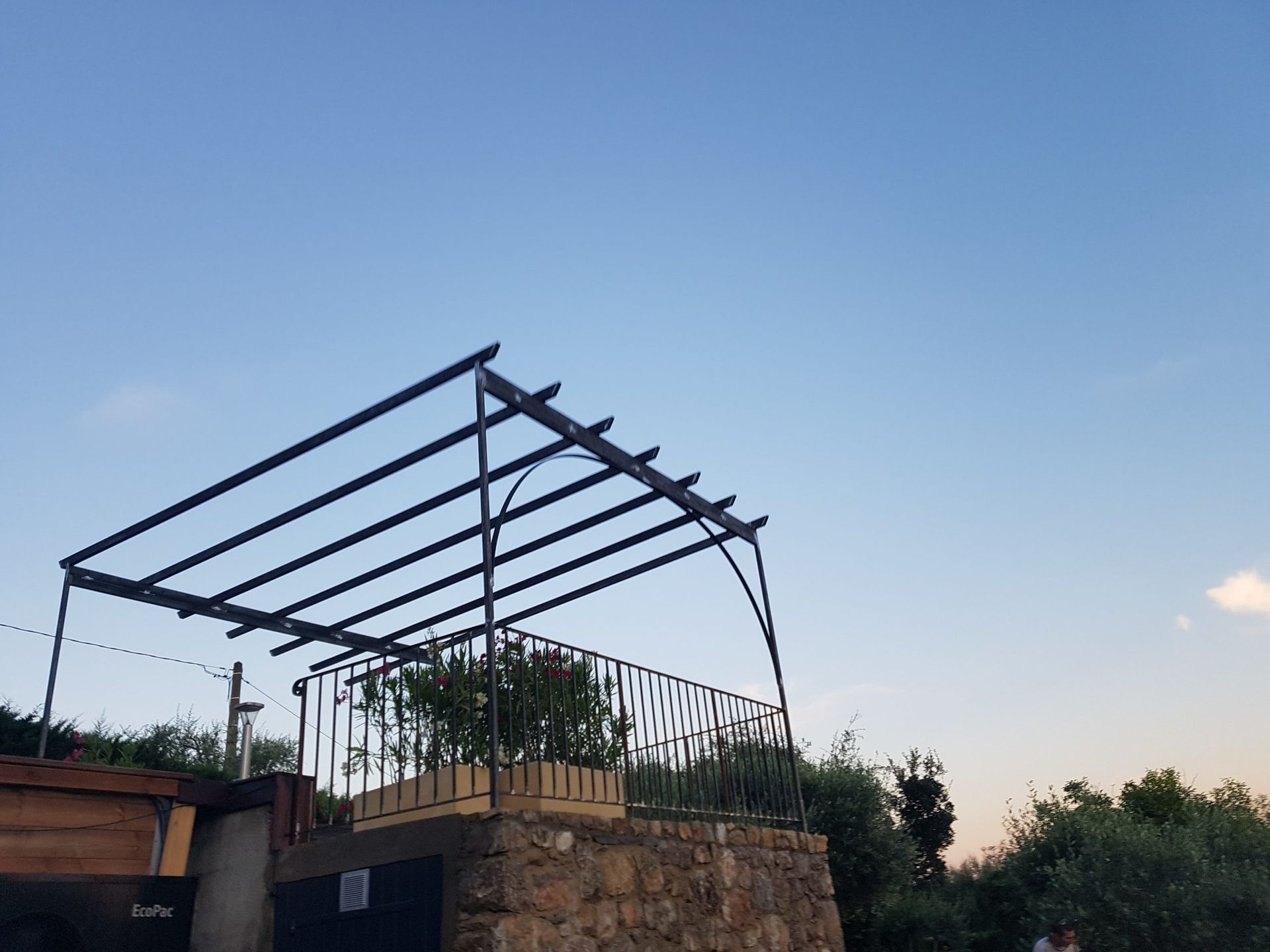 Charpente métallique partiellement construite au-dessus d'une petite structure au coucher du soleil, avec des arbres et un ciel bleu en arrière-plan.