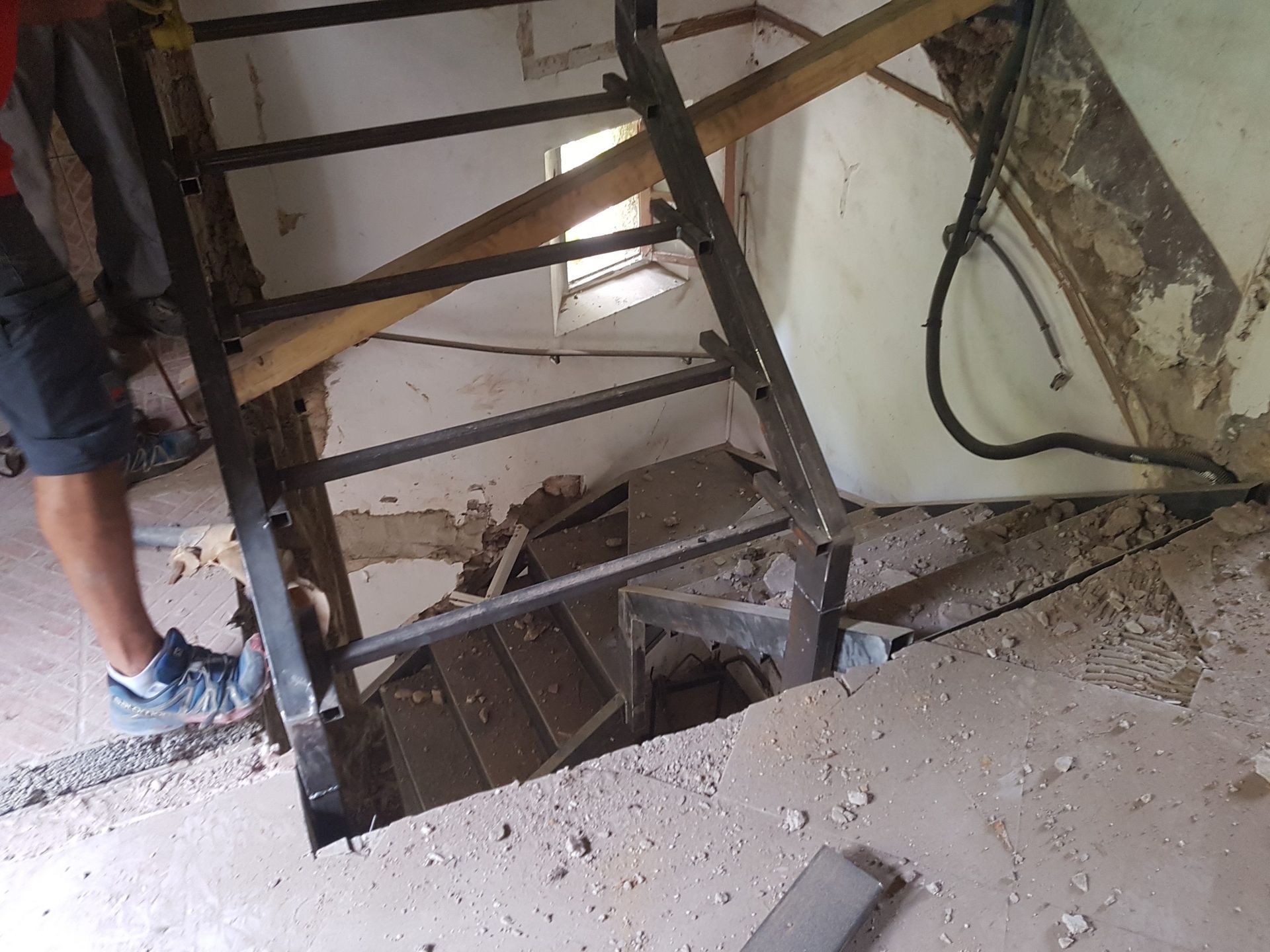 Vue plongeante sur un escalier intérieur raide, avec des poutres apparentes et les jambes d'une personne visibles à gauche.