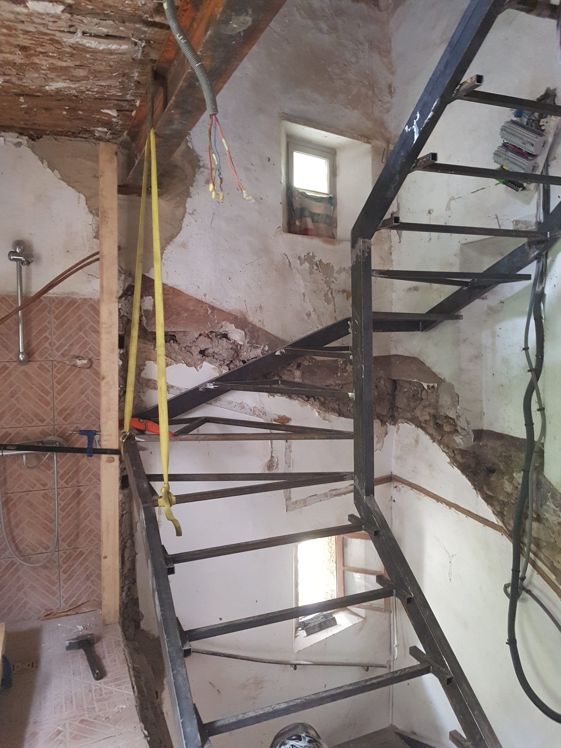 Intérieur de la cage d'escalier endommagé avec des fils électriques apparents, des murs fissurés et un escalier en métal noir