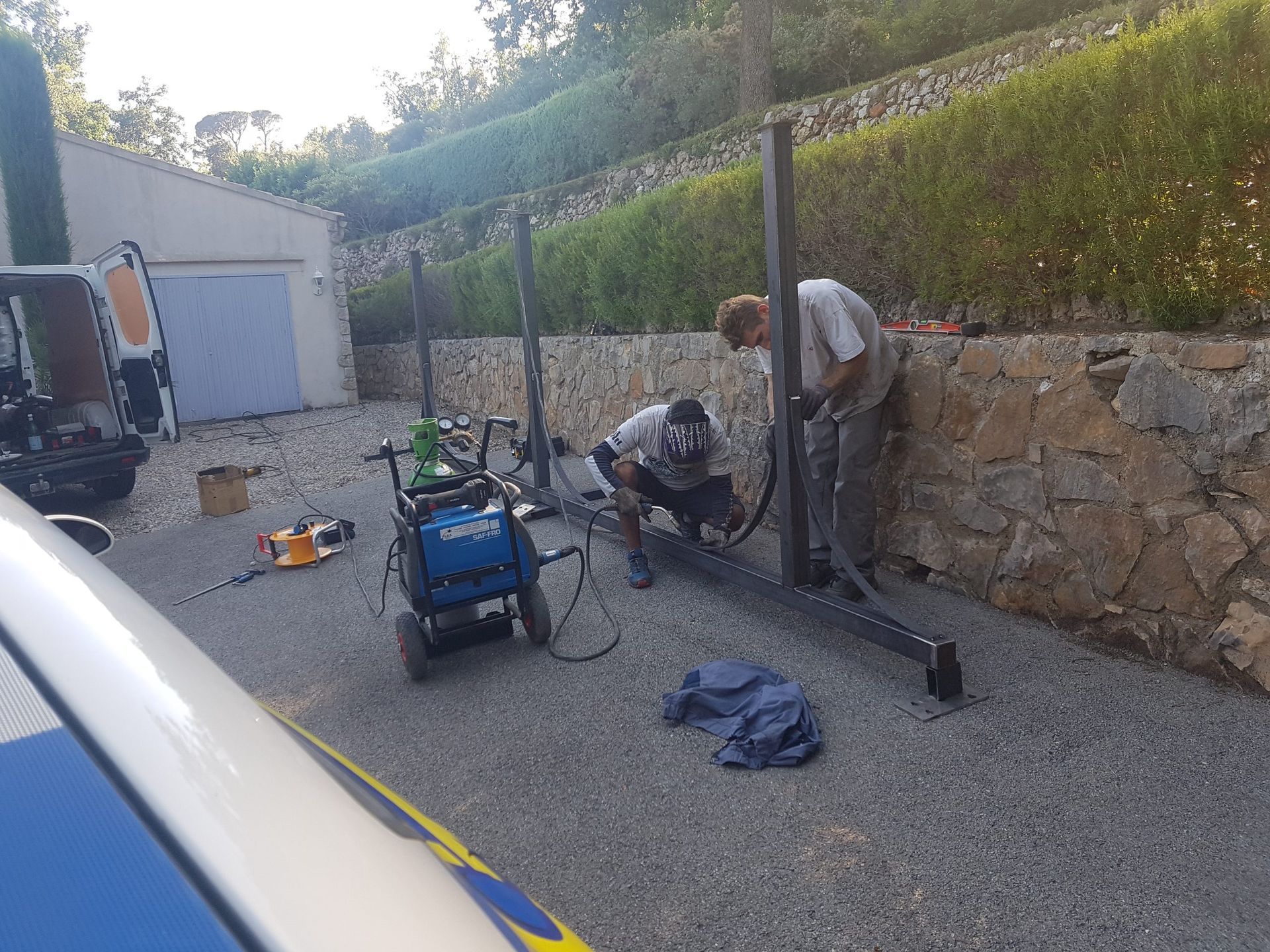Des personnes travaillent avec des outils près d'un muret en pierre, dans une allée de gravier, avec une machine bleue et des câbles.