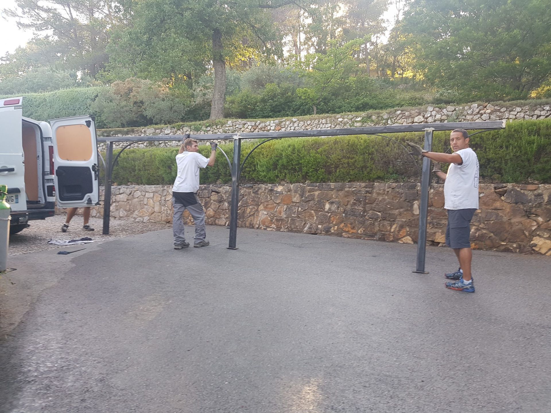 Trois personnes en chemises blanches tiennent une longue perche à côté d'un camion dans une allée près d'un mur de pierre.