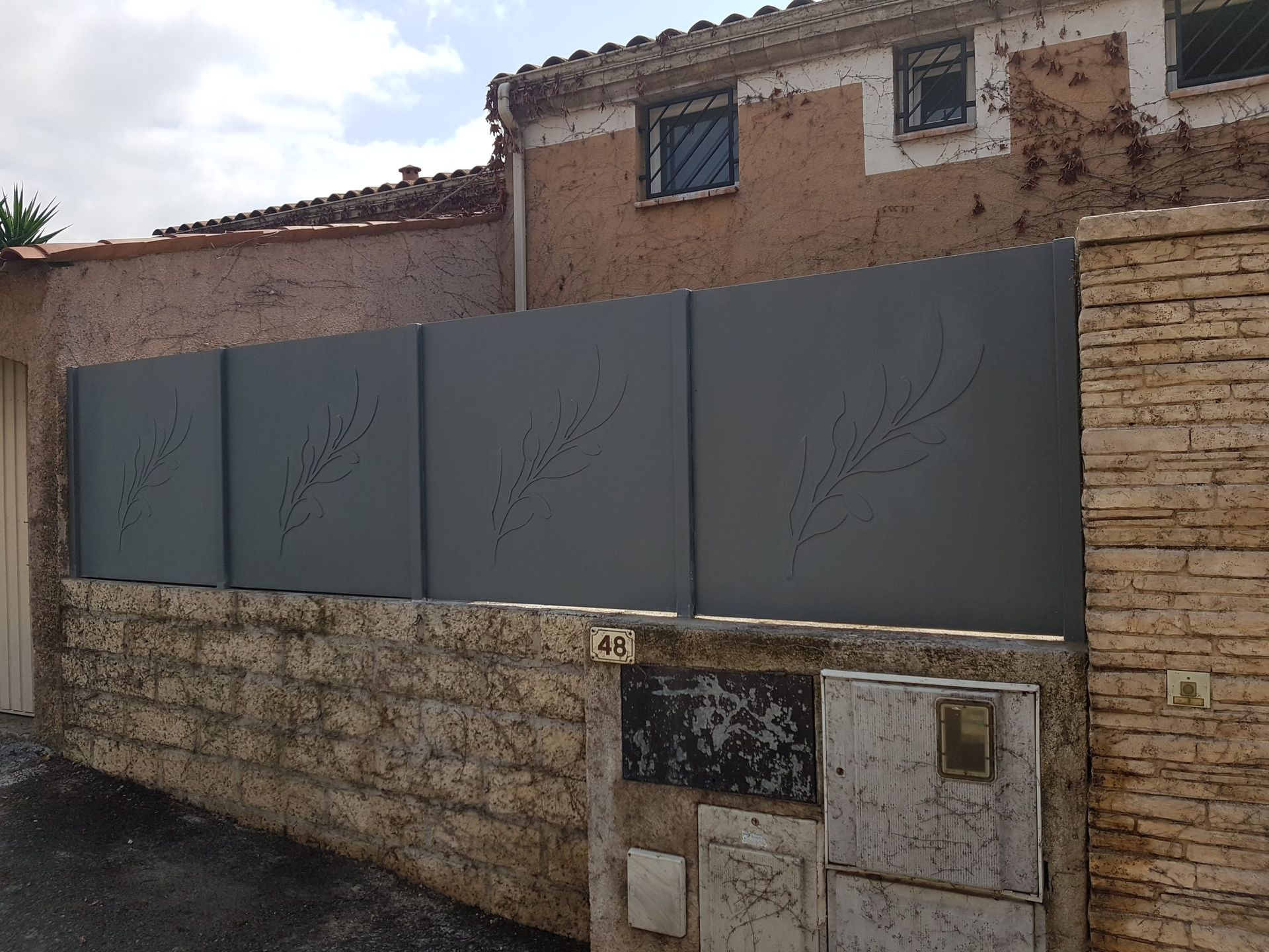 Maison en construction ou en réparation, avec une longue clôture métallique grise et un mur en pierre.