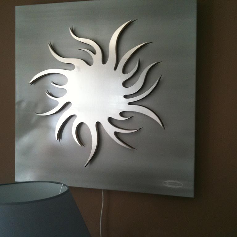 Sculpture murale argentée en forme de soleil sur un mur gris, au-dessus d'un abat-jour.