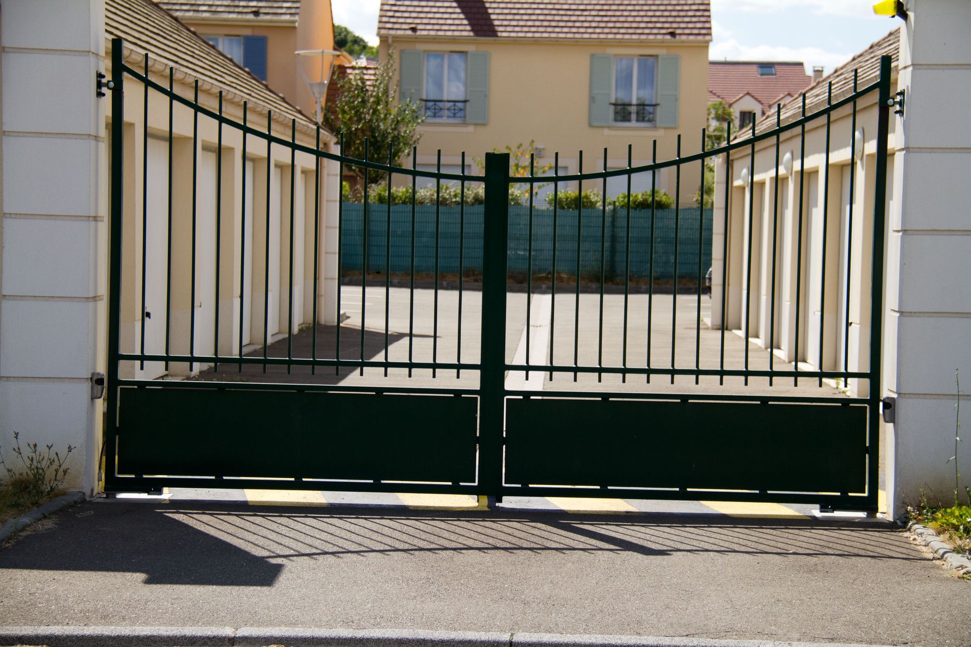 Un portail métallique vert fermé, entre des murs blancs, bloque l'accès à une allée devant des maisons.
