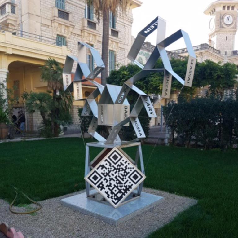 Sculpture abstraite en métal, installée en extérieur dans une cour paysagée et dotée d'un panneau à code QR.