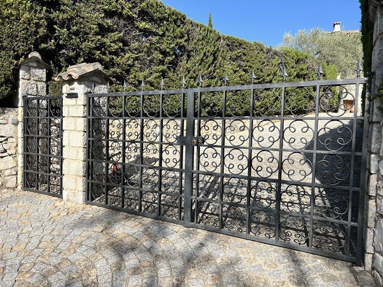 Un portail en fer forgé noir se dresse devant un mur de pierre et une haie recouverte de lierre, avec un premier plan pavé.