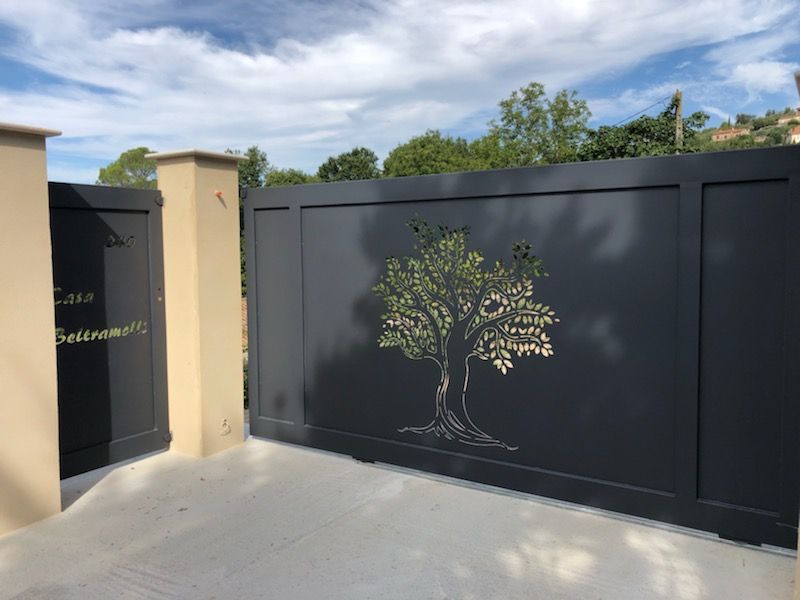 Portail d'allée noir orné d'un motif d'arbre peint, adossé à un mur beige sous un ciel bleu.