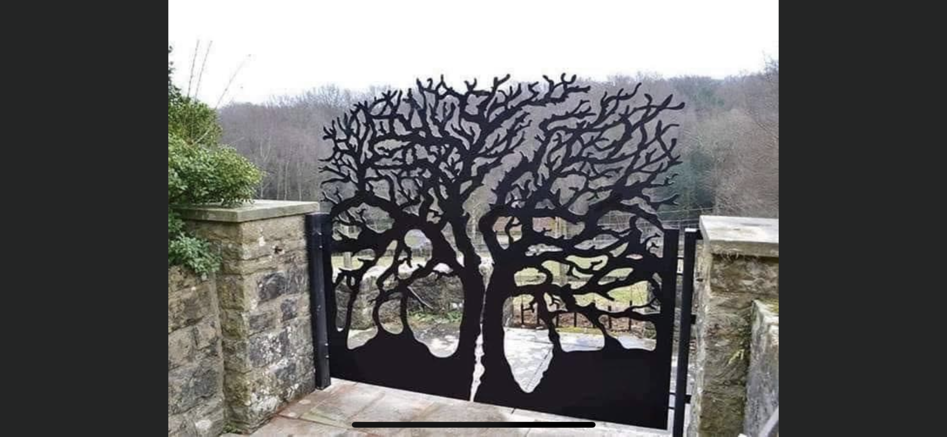 Portail en métal noir en forme d'arbre, encadré par des piliers de pierre, avec un paysage boisé en arrière-plan.