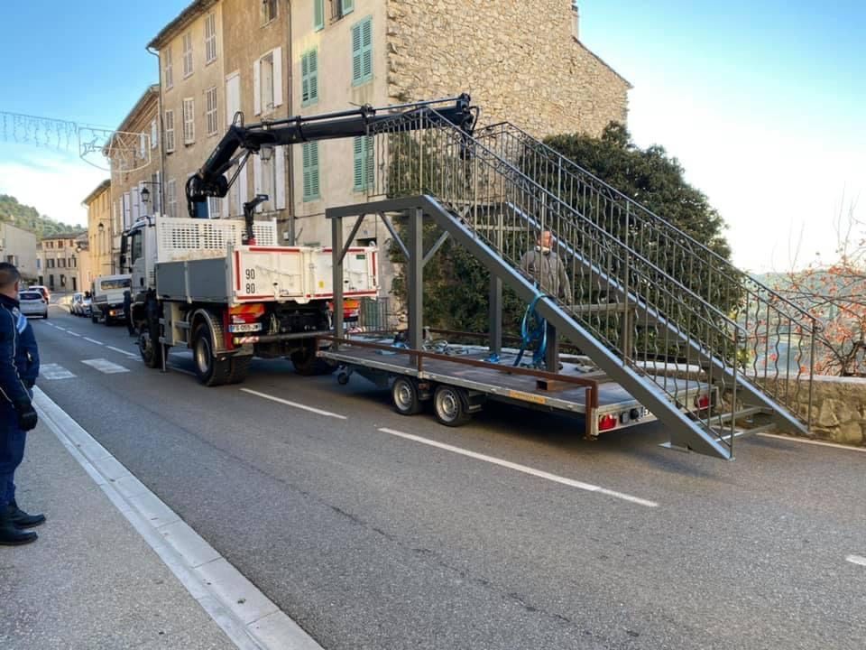 Une grue montée sur camion soulève un grand escalier métallique à côté d'un bâtiment dans une rue de la ville.