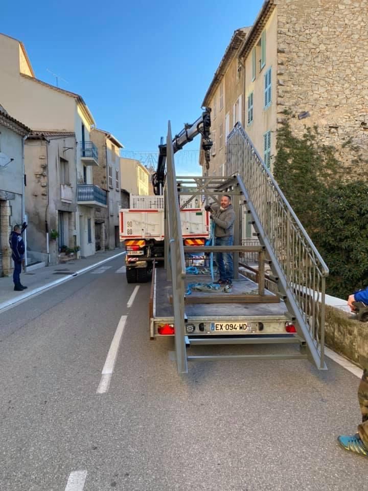 Scène de rue avec un camion et une structure d'escalier métallique bloquant une route étroite entre des bâtiments en pierre