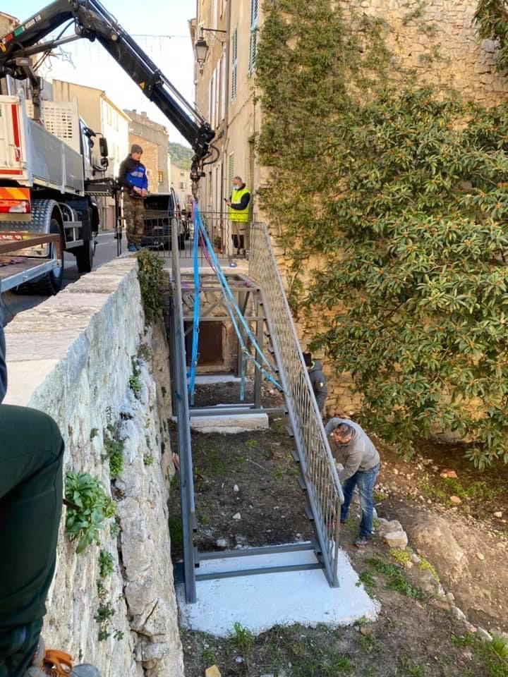 Des ouvriers utilisent une grue pour installer un escalier métallique à côté d'un mur de pierre et d'une haie.