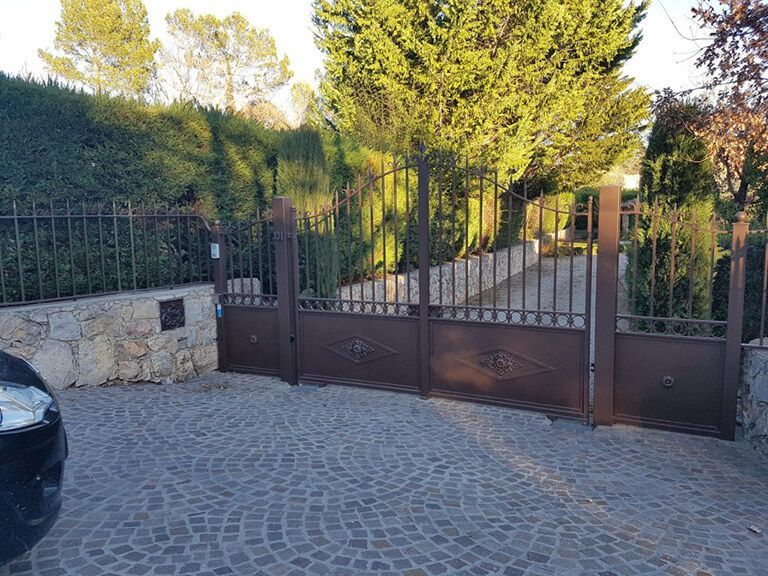 Allée pavée avec un portail en métal, un mur en pierre et de grands arbres derrière.