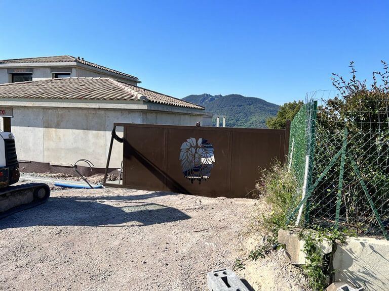 Portail en métal marron avec découpe circulaire à côté d'une maison sur une allée de gravier, montagnes en arrière-plan