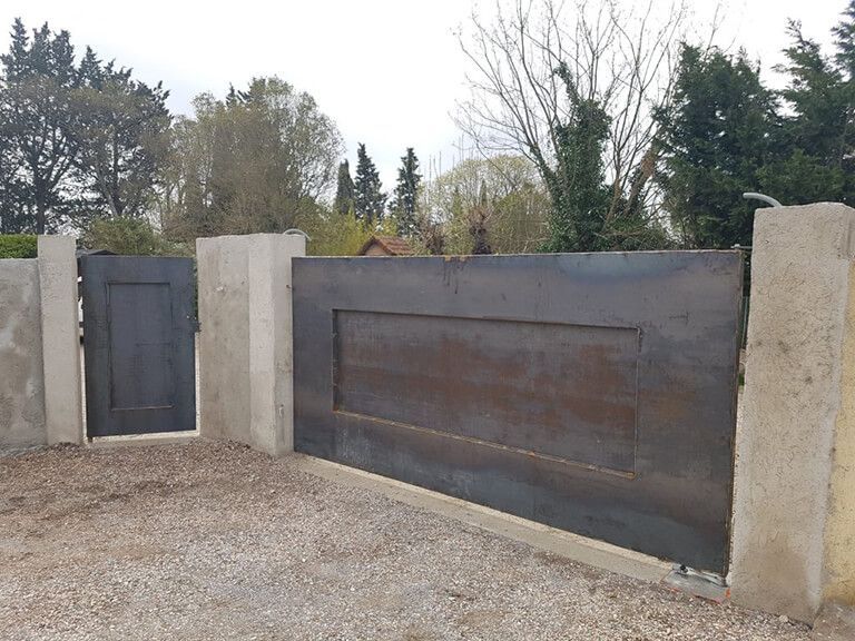 Portail métallique moderne installé sur des poteaux en béton le long d'une allée de gravier, avec des arbres en arrière-plan.