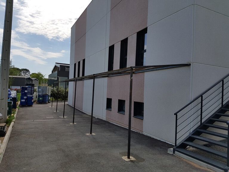 Bâtiment blanc moderne avec escalier, fenêtres étroites et parking pavé avec poteaux et poubelles.