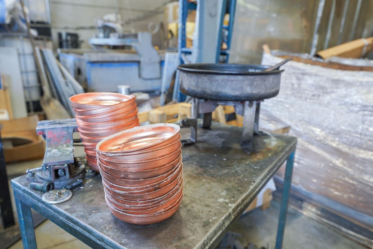 Des casseroles en cuivre sur un établi dans un atelier, avec une grande bouilloire en métal et des équipements industriels en arrière-plan.