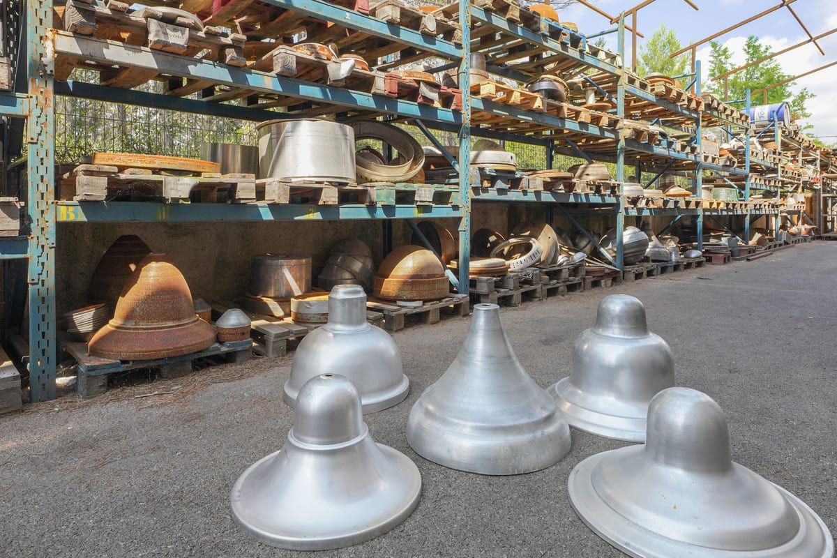 Des cloches industrielles argentées posées au sol devant des étagères métalliques remplies d'autres cloches, à l'extérieur.