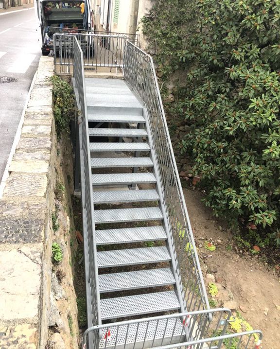 Escalier extérieur en métal à forte pente avec rampes, jouxtant un mur de pierre et une végétation dense.
