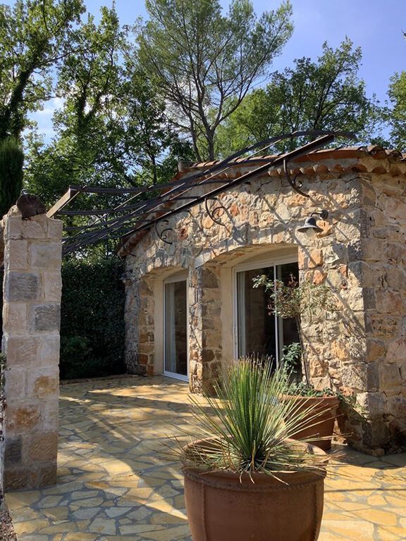 Maisonnette en pierre avec porte cintrée, patio ensoleillé et plante en pot sous les arbres