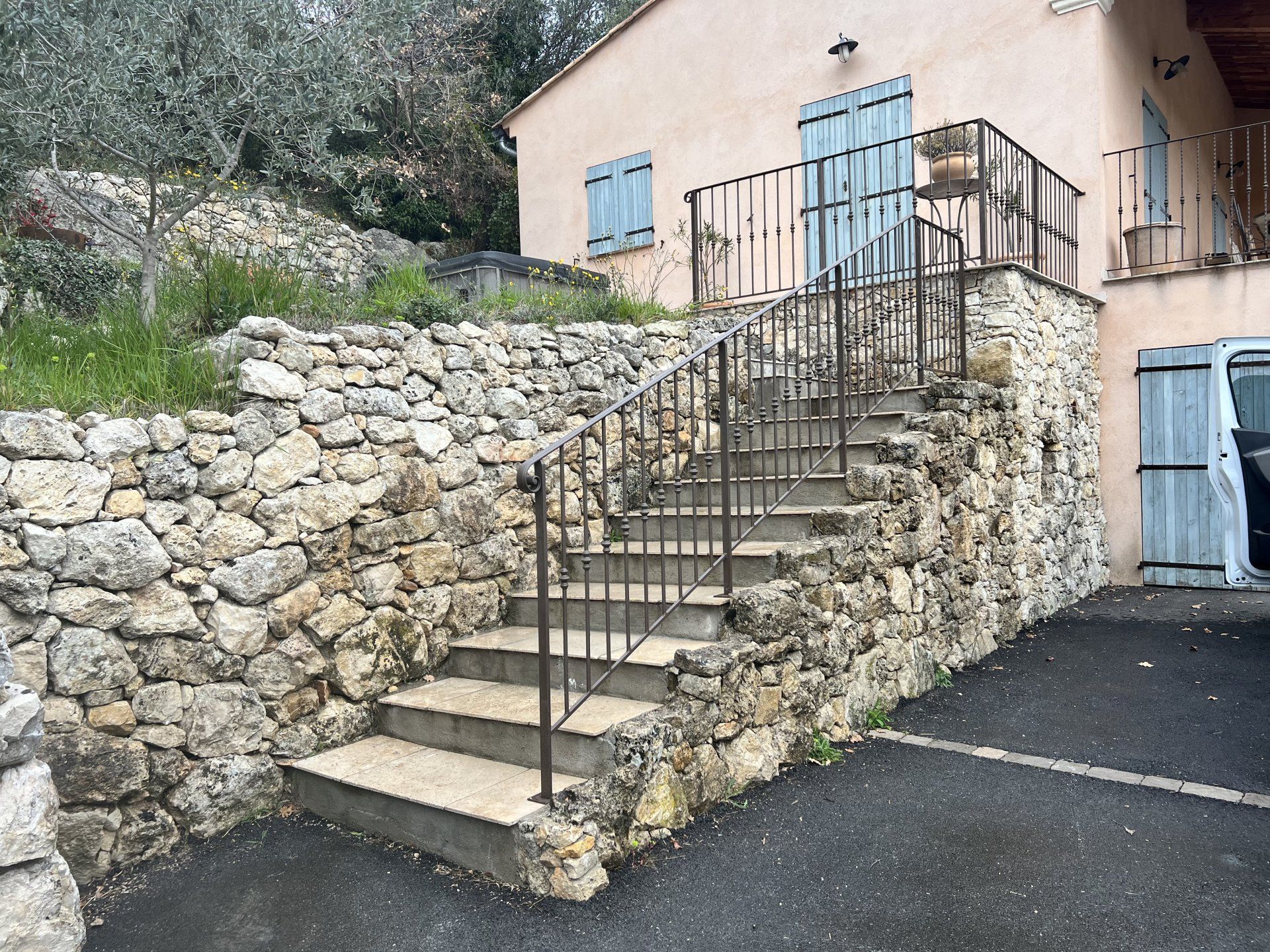 Un escalier en pierre avec une rampe en métal noir mène à une porte bleue, à côté d'une maison couleur pêche et d'un mur en pierre.