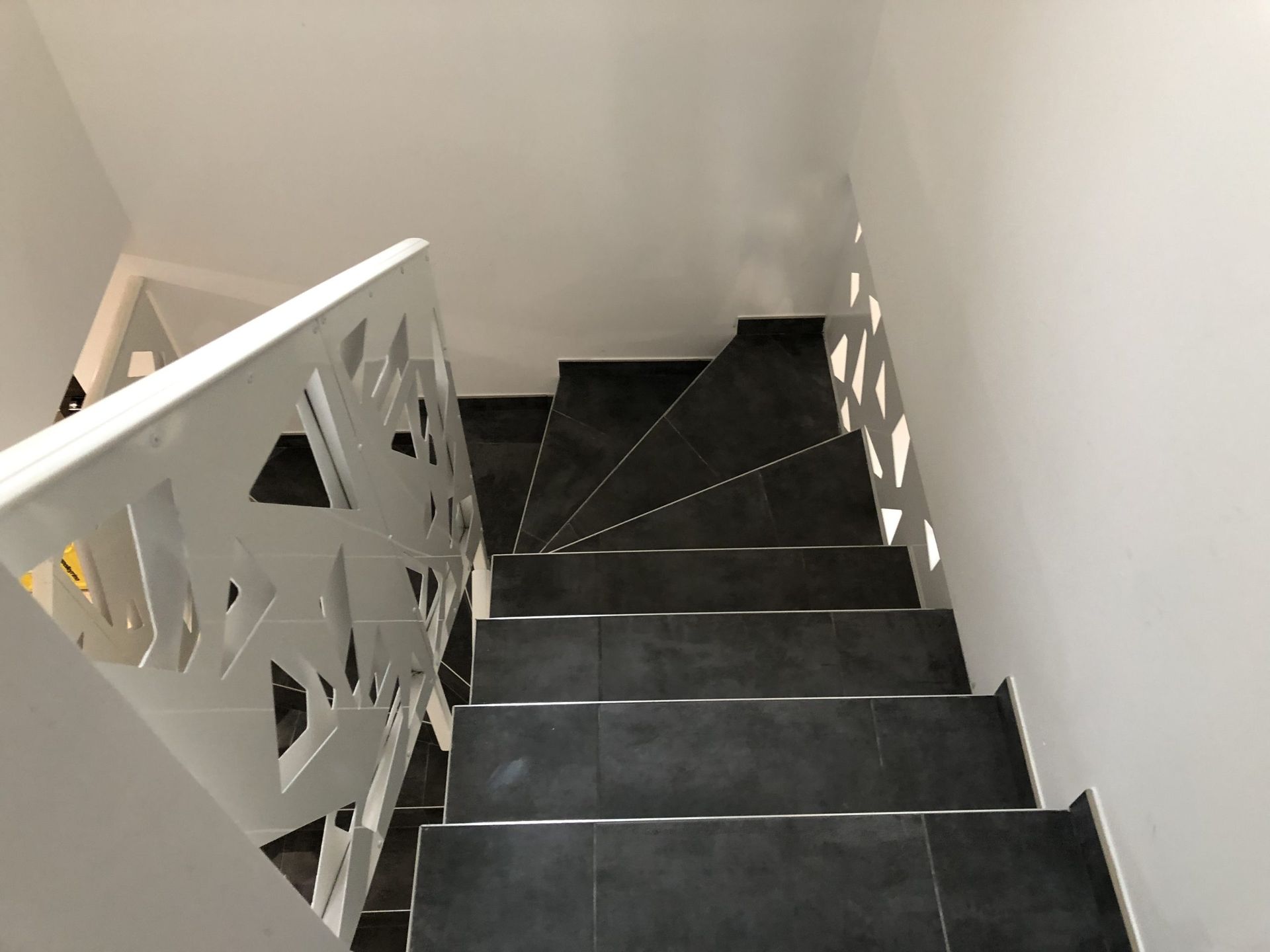 Escalier intérieur étroit avec marches sombres, murs blancs et rampe décorative blanche.