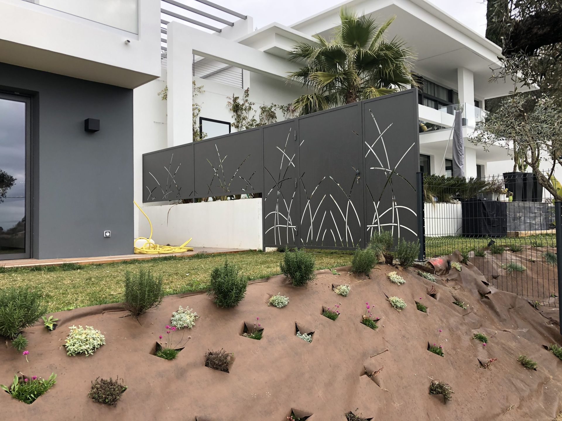 Maison moderne en construction, avec façade blanche, mur d'intimité noir et jardin paysager en pente.