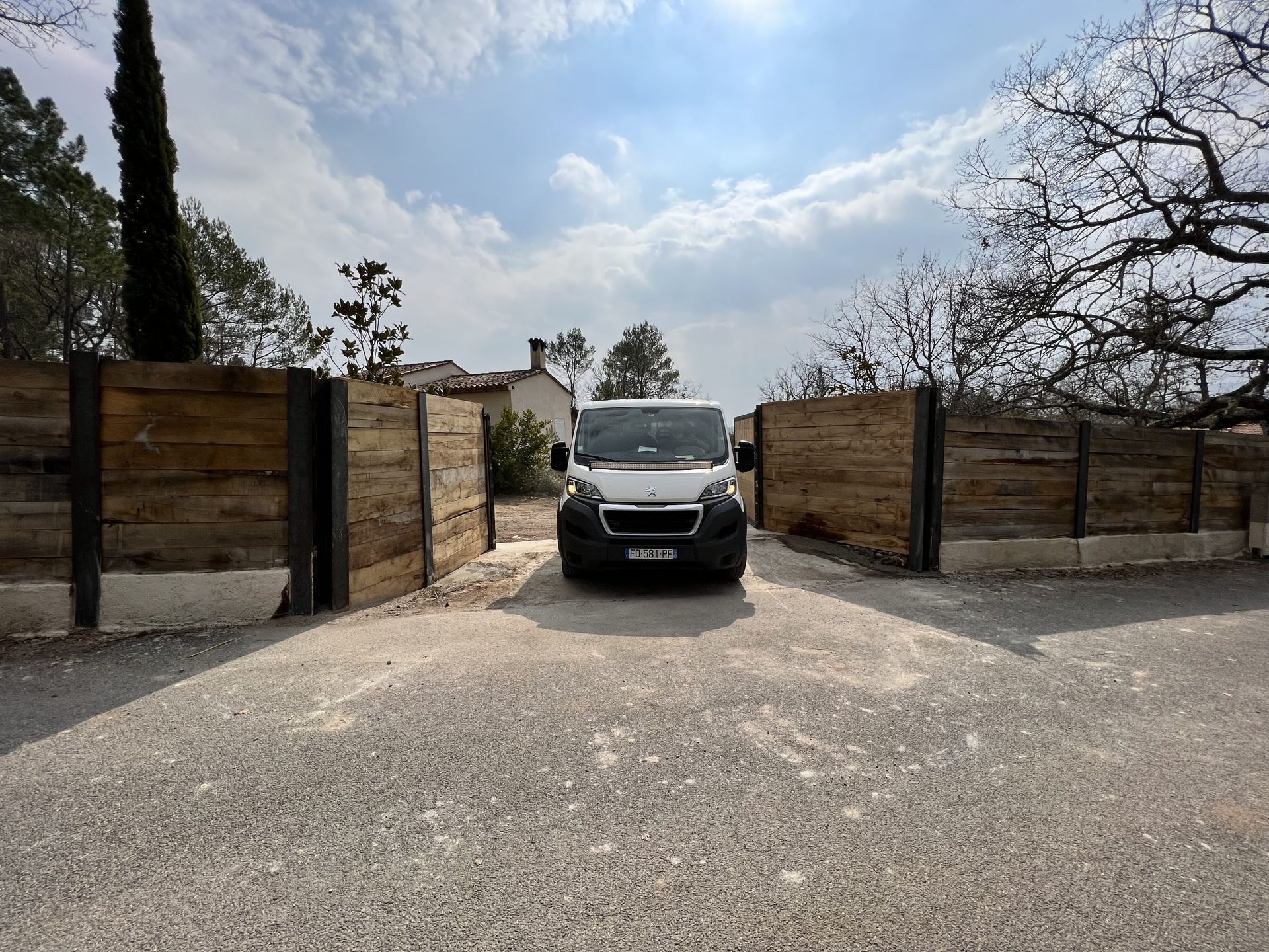 Une camionnette noire entre par un portail en bois ouvert sur une allée de gravier sous un ciel partiellement nuageux.