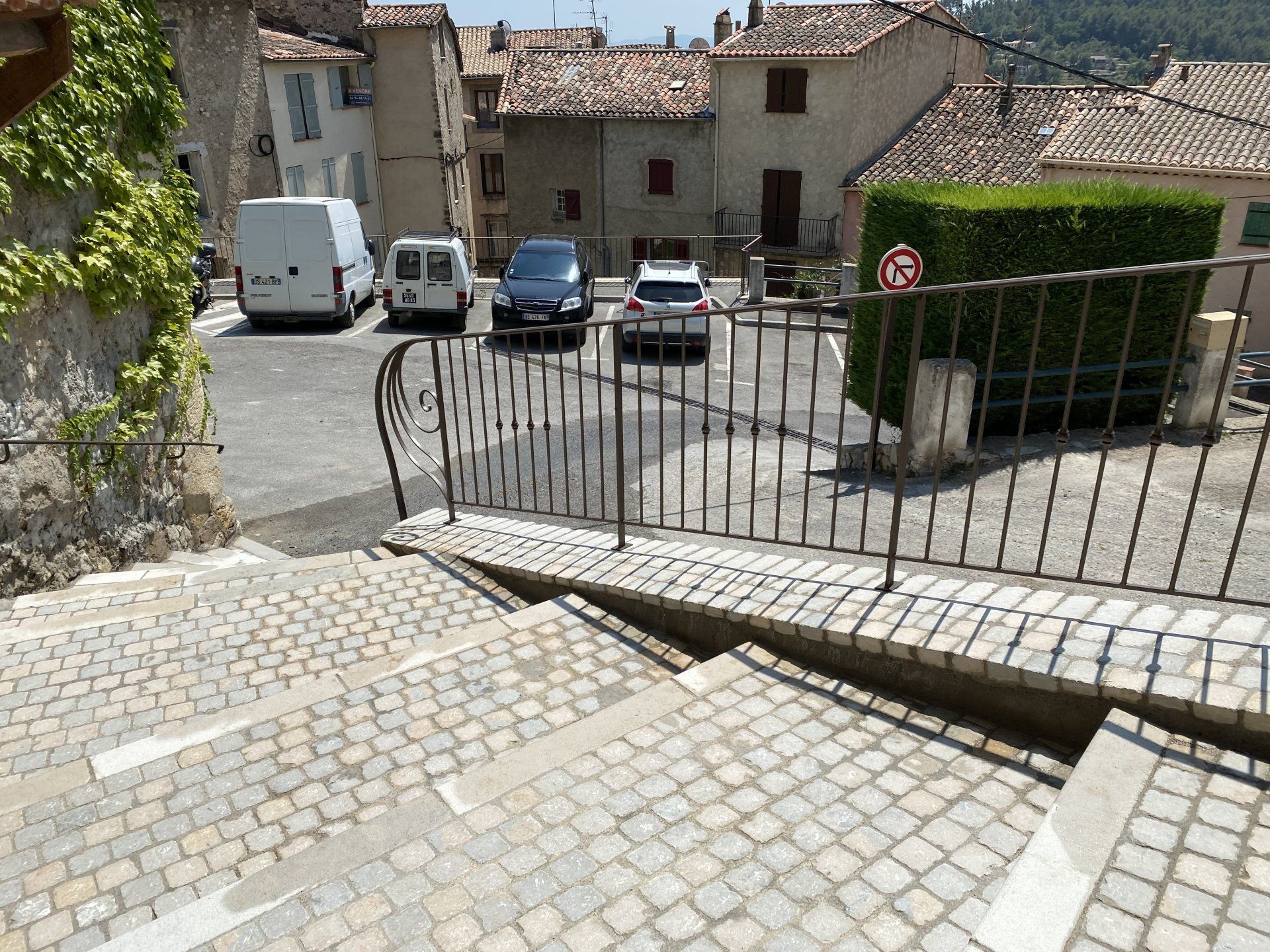 Des marches en pierre descendent vers une cour fermée par un portail, au-delà de laquelle se trouvent des voitures garées et des bâtiments du village.