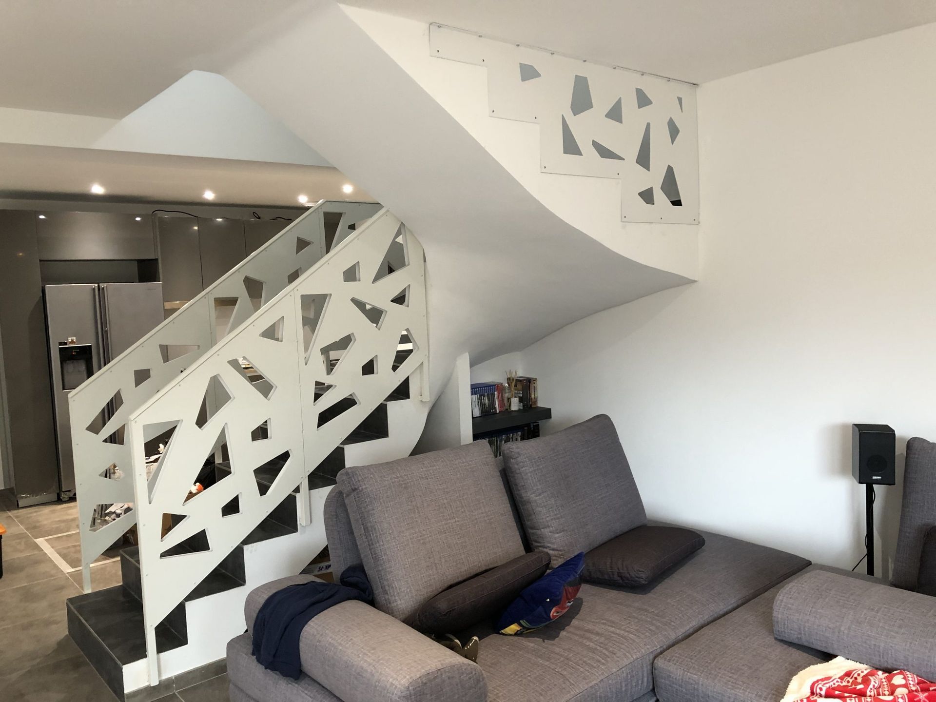 Salon moderne avec canapé d'angle gris, escalier blanc et balustrade à motifs géométriques.