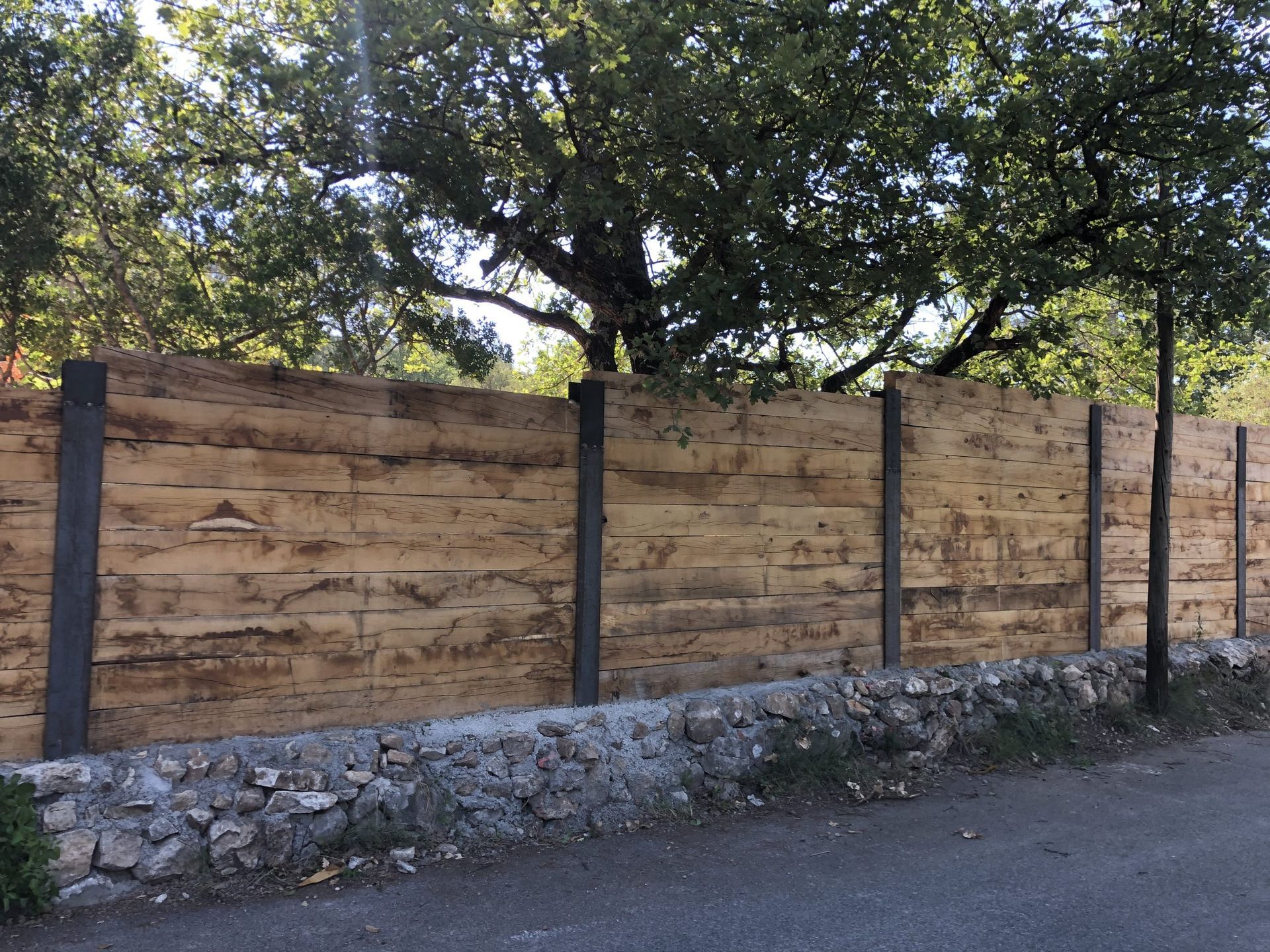 Clôture en pierre et en bois au bord d'une route, avec des arbres feuillus formant une voûte au-dessus.