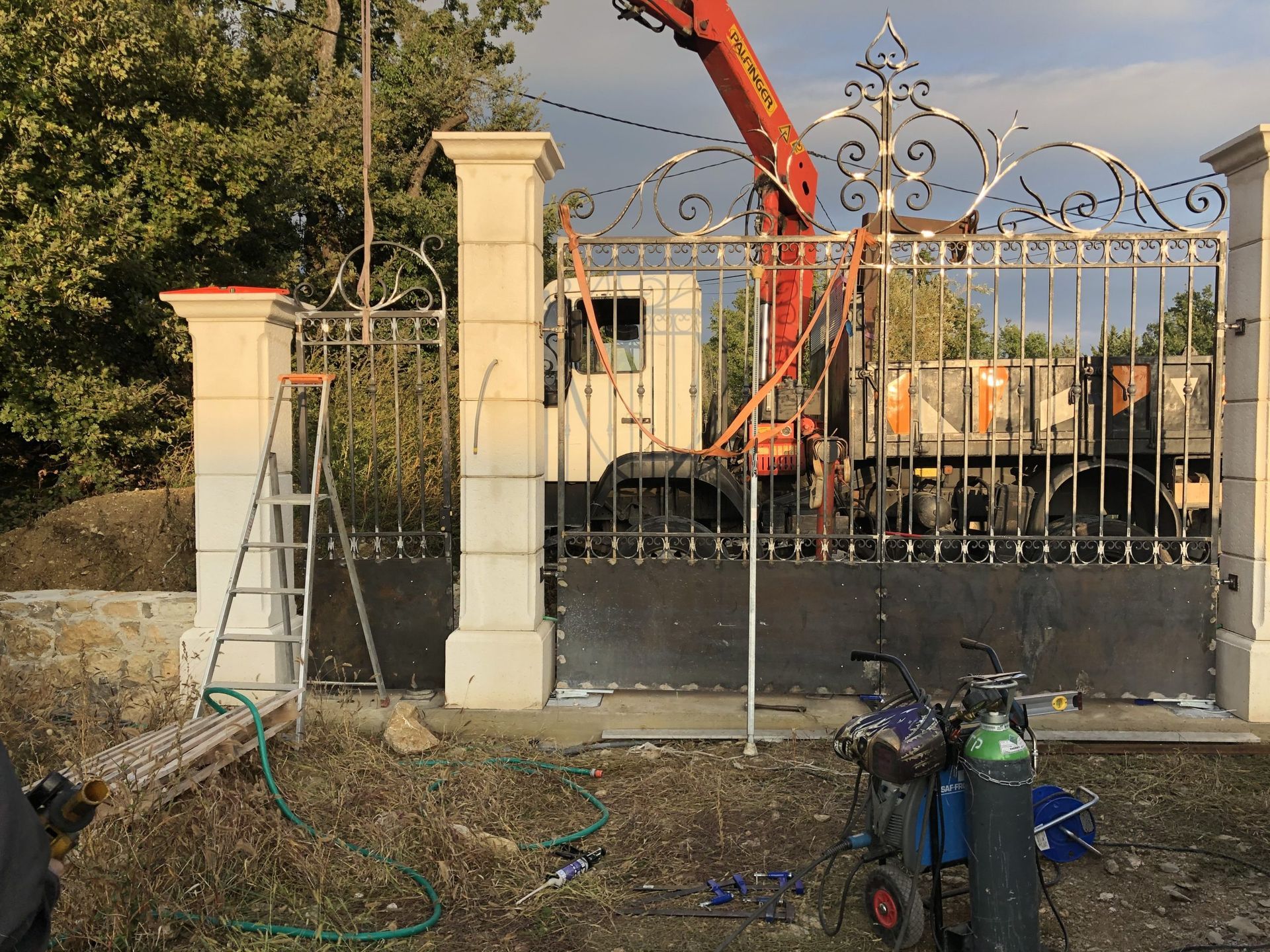 Chantier de construction avec portail métallique orné, piliers en béton et une grue rouge derrière la clôture.