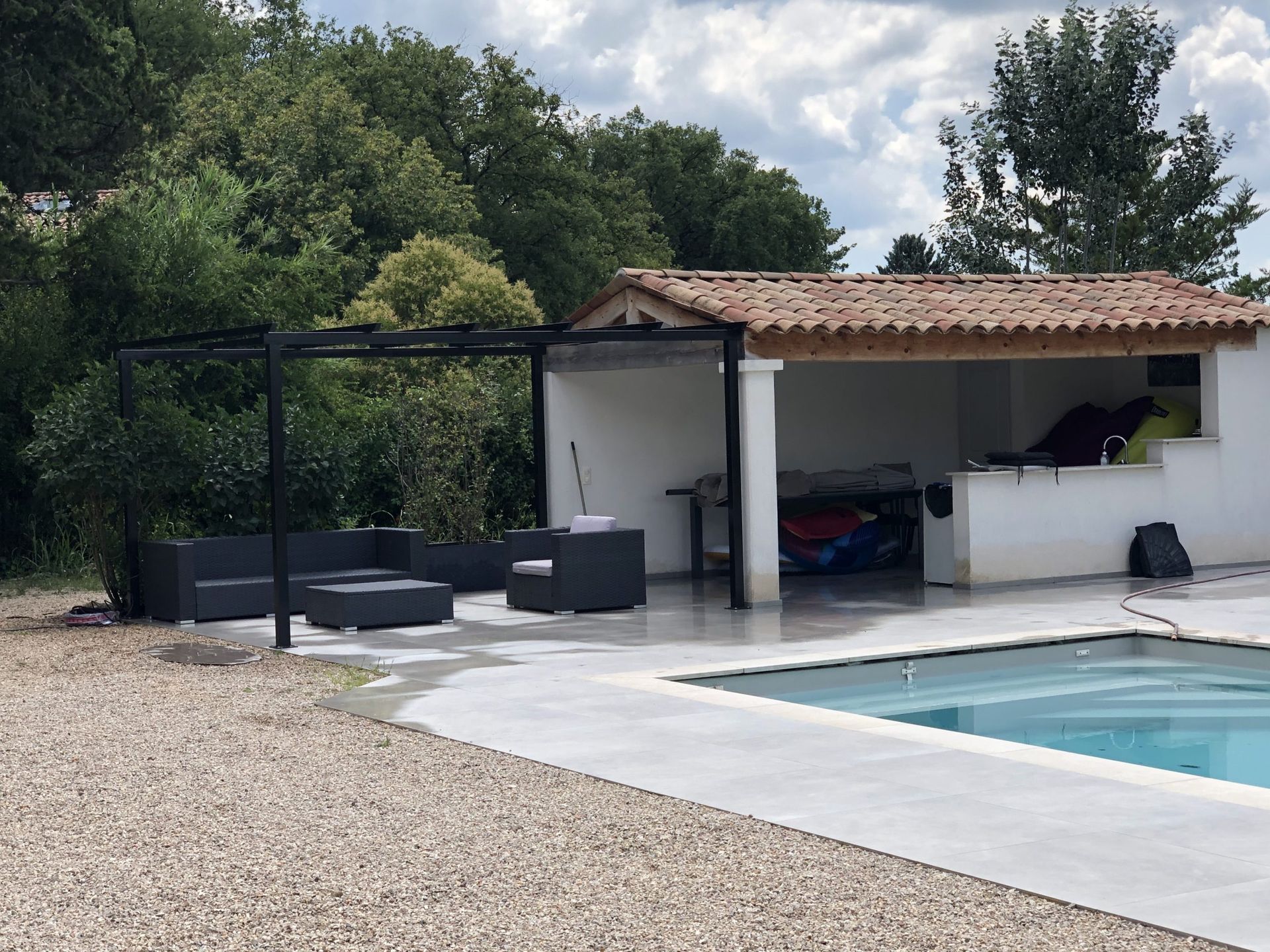 Terrasse au bord de la piscine avec coin salon couvert, terrasse en béton blanc, gravier et arbres sous un ciel nuageux