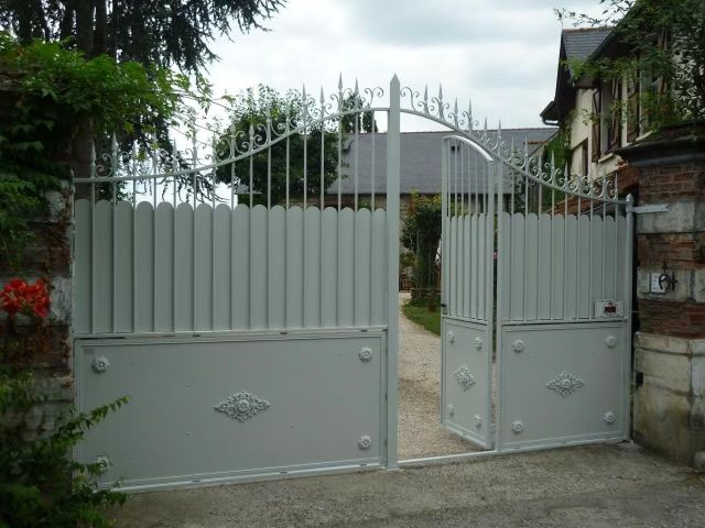 Un portail métallique gris fermé marque l'entrée d'une maison, avec un étroit passage et de la verdure derrière.