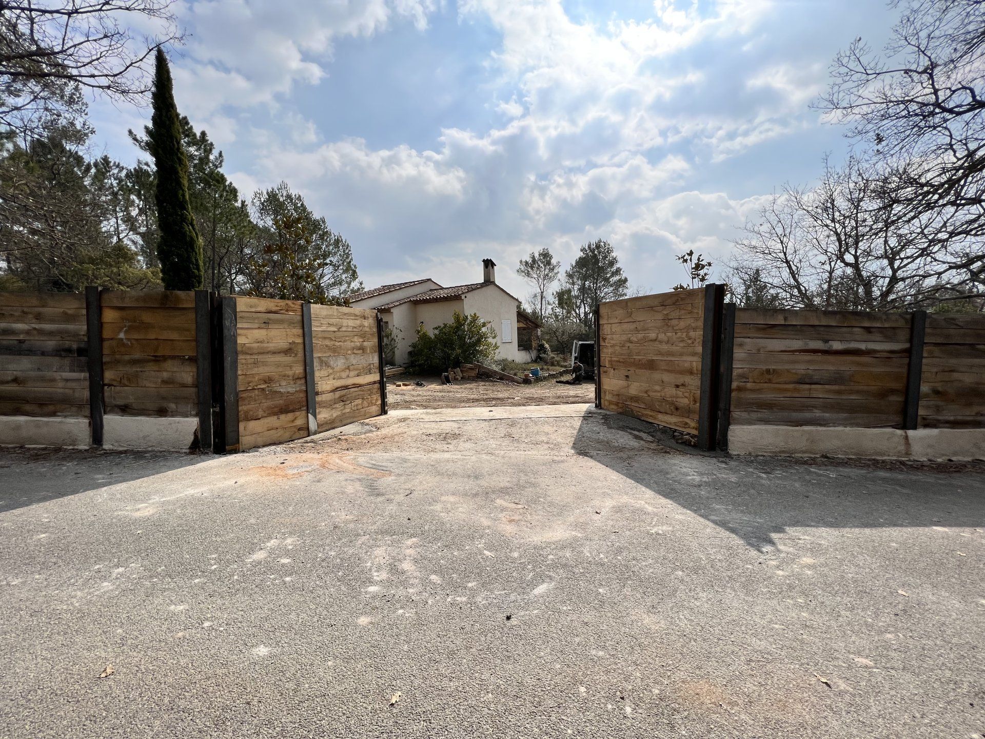 Des portails en bois ouverts mènent à une allée de gravier et à une maison sous un ciel nuageux.