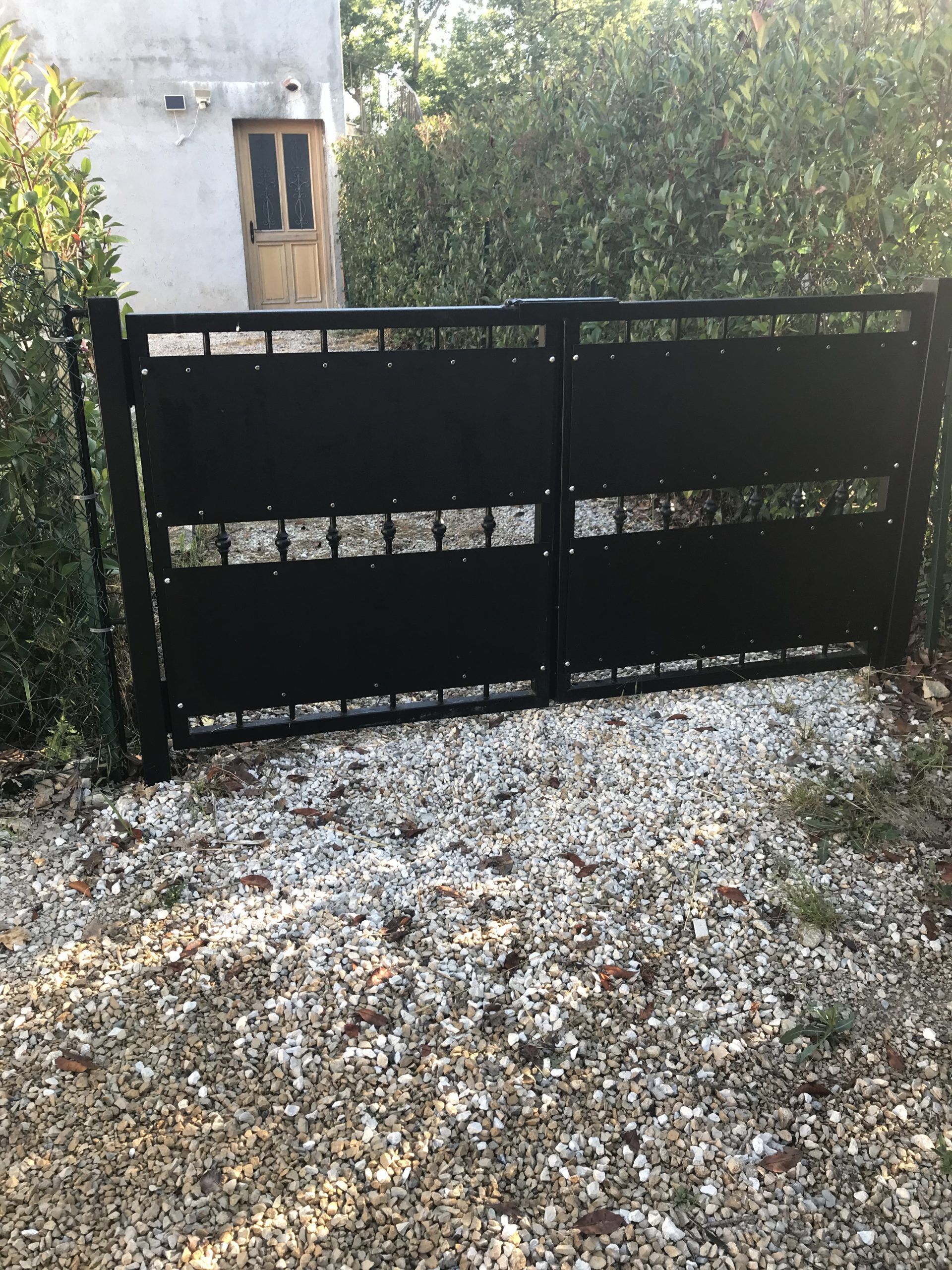 Portail d'allée en métal noir devant une maison aux murs blancs, avec des arbres à l'arrière et un sol en gravier au premier plan.