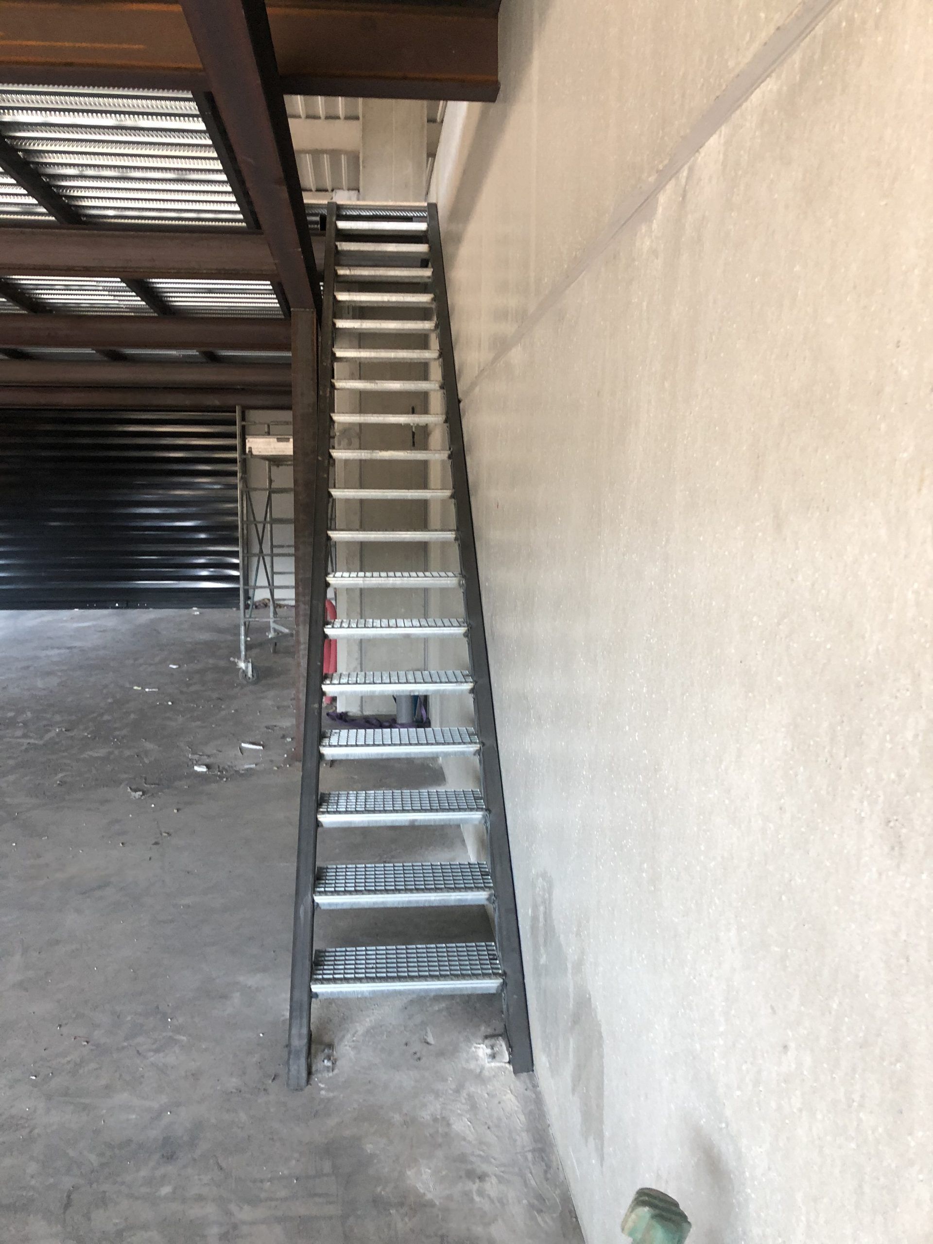 Escalier métallique étroit contre un mur beige sous une structure surélevée dans un espace industriel