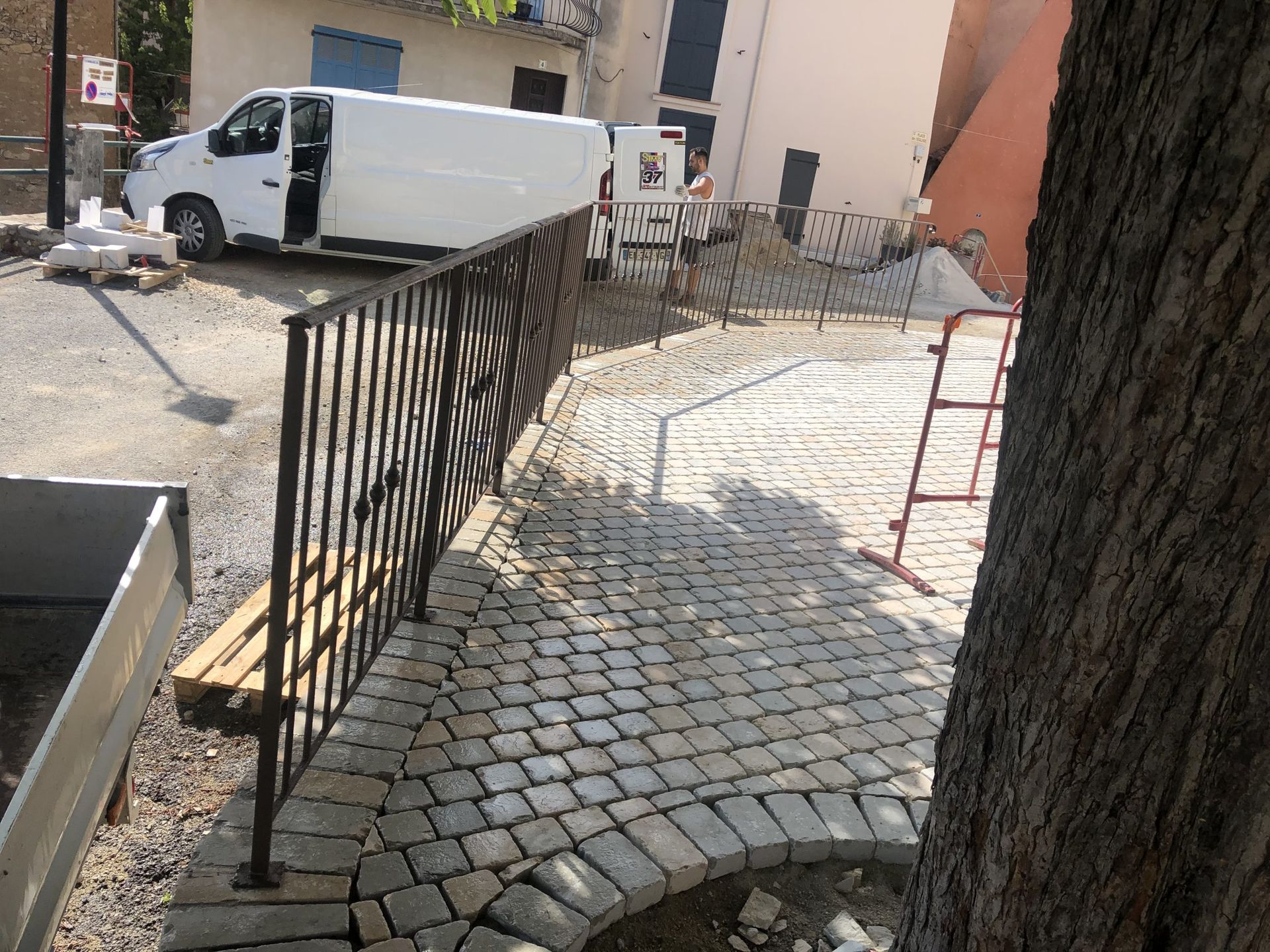 Allée pavée longeant une rambarde noire, avec une camionnette blanche garée près d'un bâtiment et d'un tronc d'arbre au premier plan.