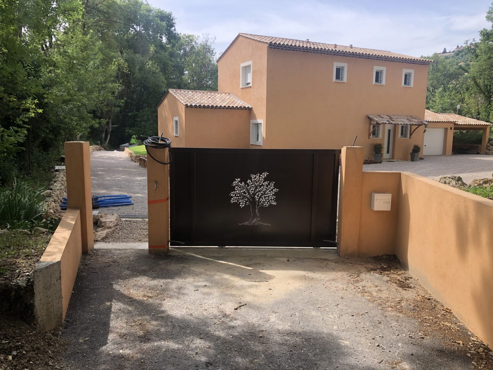 Portail ouvert donnant sur une maison beige avec une allée de gravier et des arbres à proximité