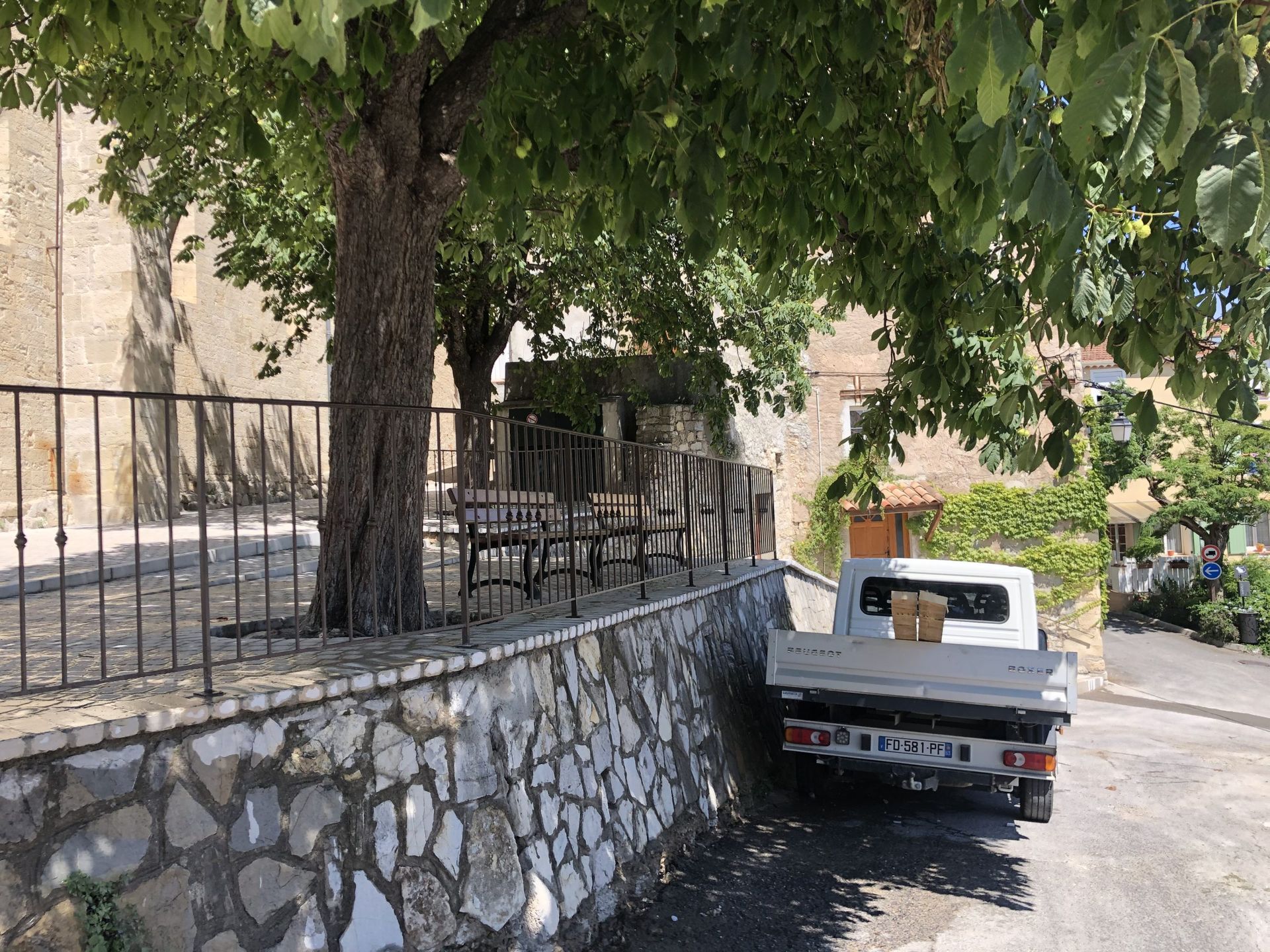 Chariot utilitaire blanc stationné sous un arbre, près d'un muret en pierre et d'une clôture métallique, dans une rue étroite.