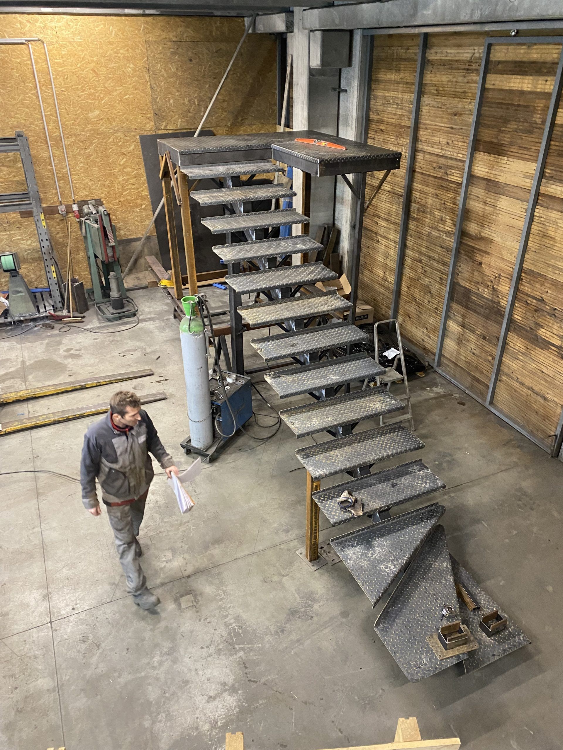 Atelier industriel avec un escalier central et une mezzanine, et une personne qui marche à l'étage inférieur.