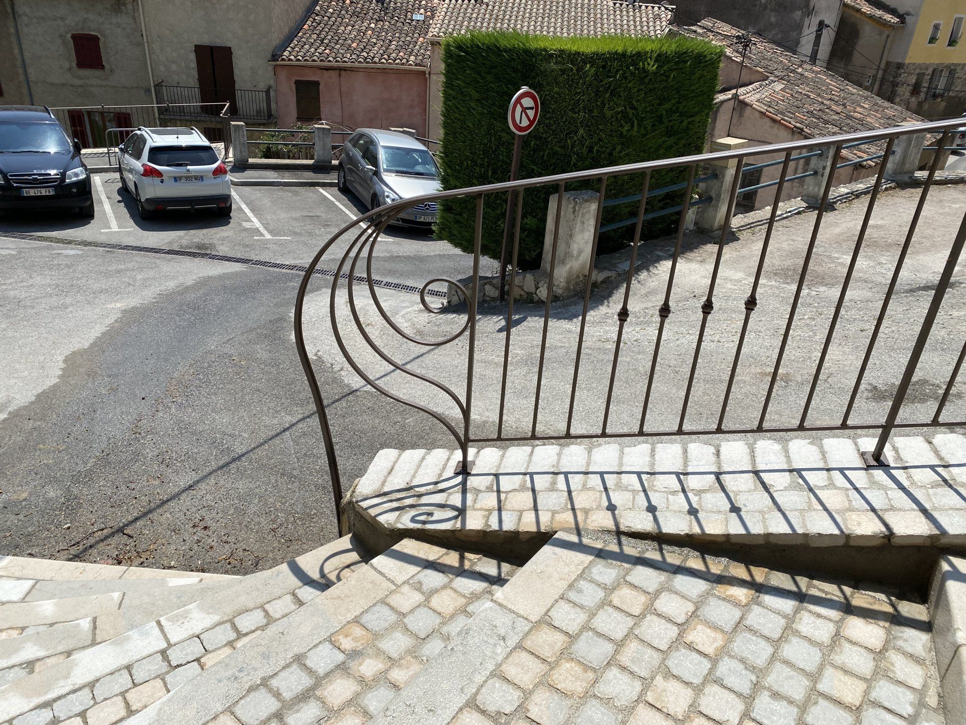 Allée pavée escarpée avec rambarde en métal noir descendant vers une rue résidentielle et des voitures garées.