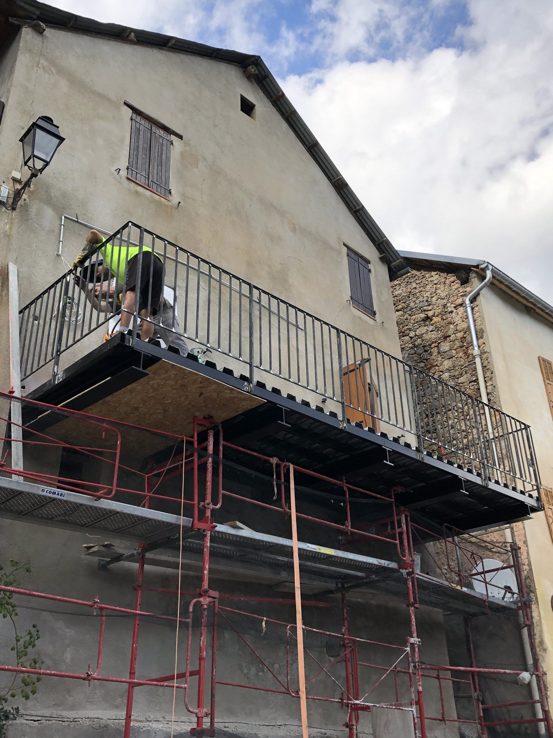 Un ouvrier sur un échafaudage répare l'extérieur d'une maison blanche avec un balcon métallique et des poteaux de soutien rouges.
