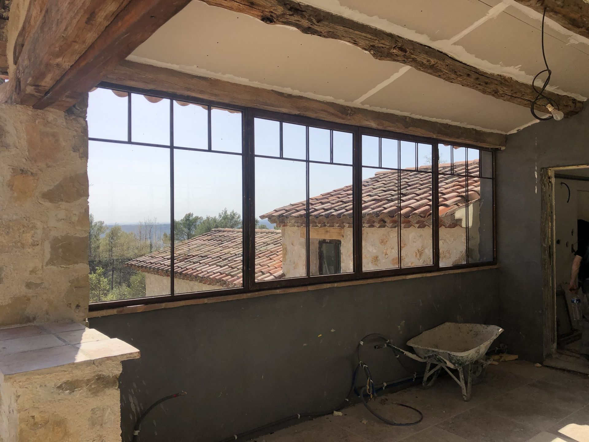 Chambre partiellement rénovée avec une grande fenêtre donnant sur les toits et une brouette au sol