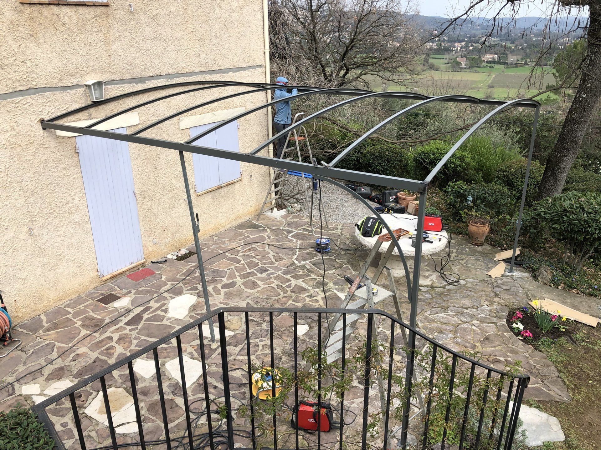 Une pergola métallique vide se dresse à côté d'une maison, avec des panneaux blancs suspendus et des débris de jardin à proximité.