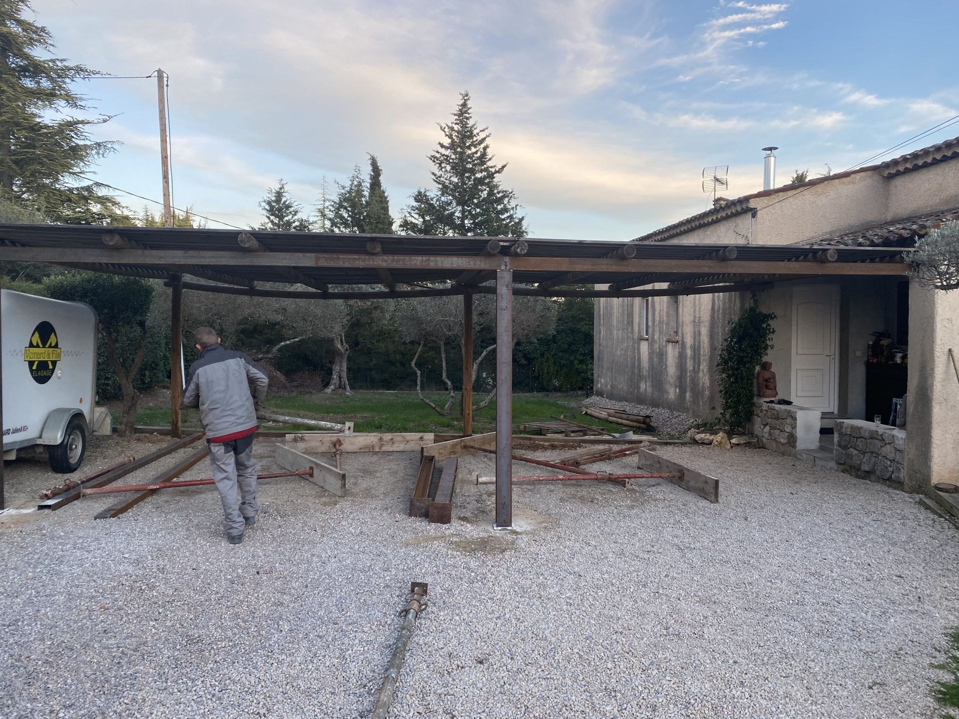 Une personne se trouve dans une cour gravillonnée, à côté d'un abri de voiture en métal et d'un petit bâtiment, avec des arbres et le ciel en arrière-plan.