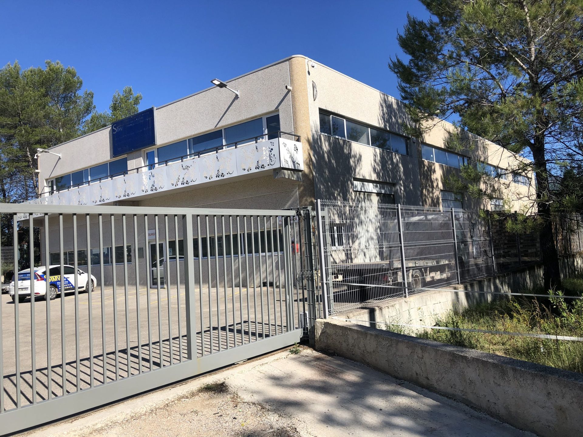 Bâtiment moderne en béton derrière un portail métallique, avec des arbres et un ciel bleu clair.