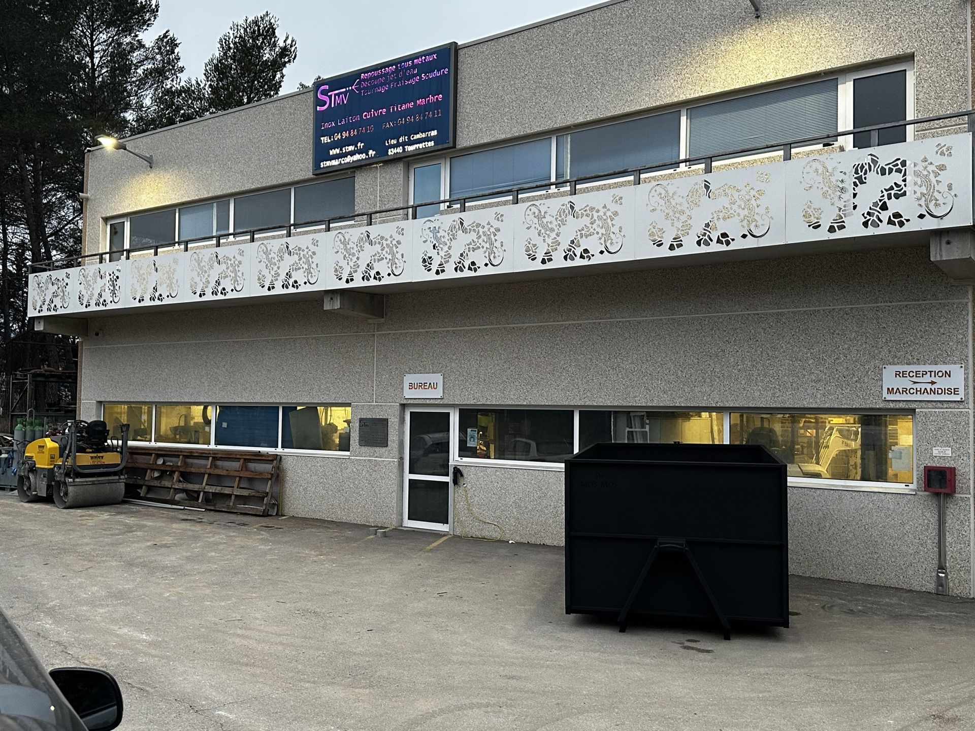 Bâtiment en béton de deux étages avec balcon couvert de graffitis, tracteur garé et grande benne à ordures noire devant.