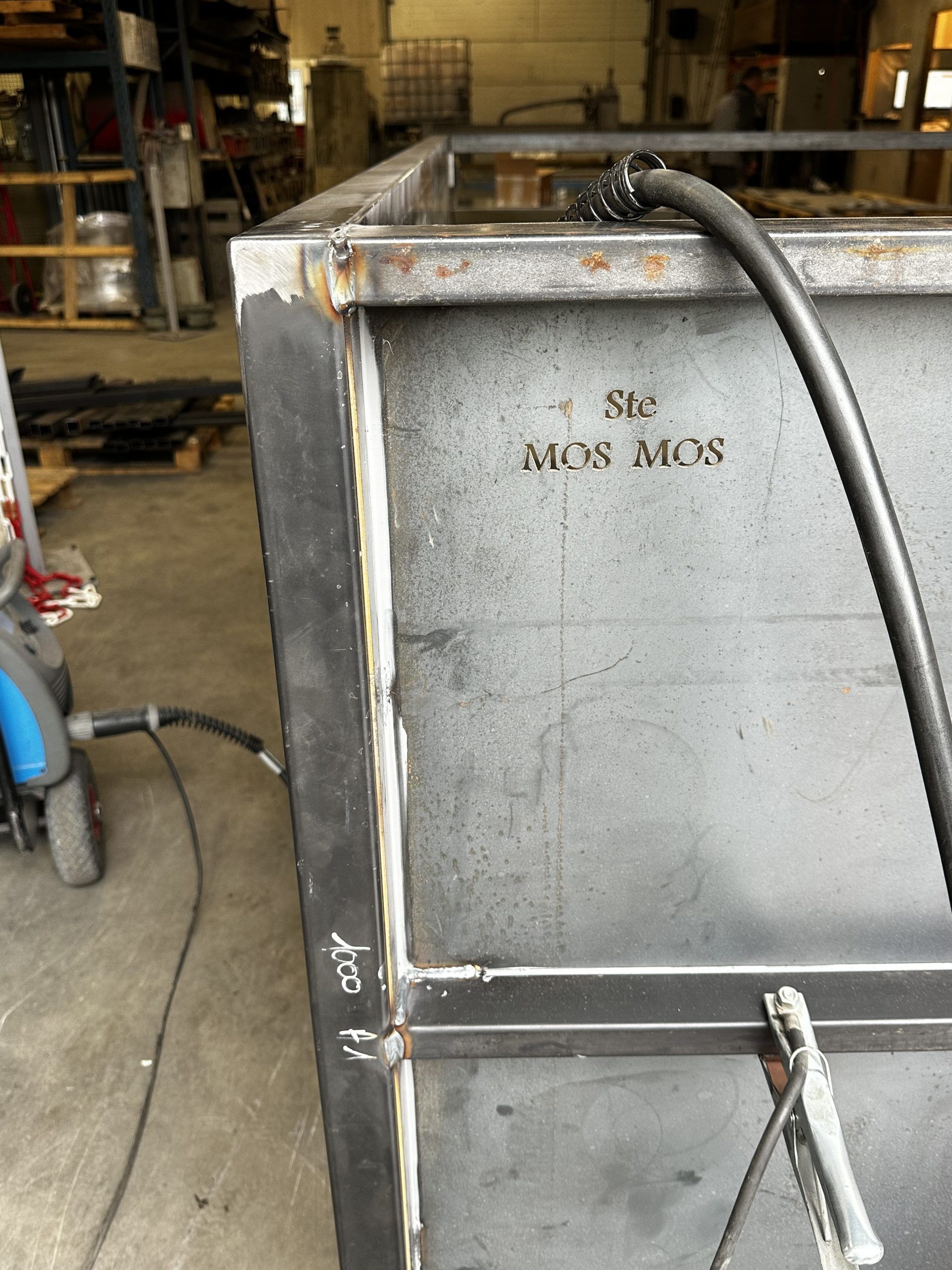 Gros plan sur une boîte industrielle en métal gris présentant des points de rouille et un tuyau d'arrosage dans un atelier.